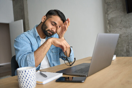 Young and tired hispanic indian student rubbing eyes and sitting at desk with a laptop.